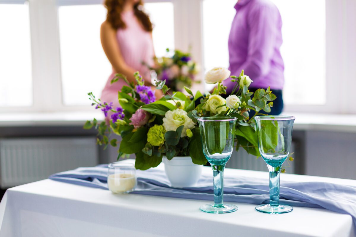 A table with two glasses and a flower arrangement with a couple in the background.